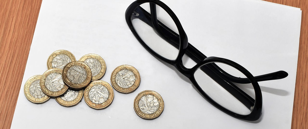A black pair of glasses lying on a table next to nine pound coins