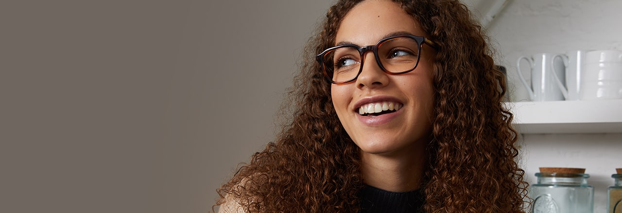 A smiling woman with long curly hair, wearing tortoiseshell glasses frames