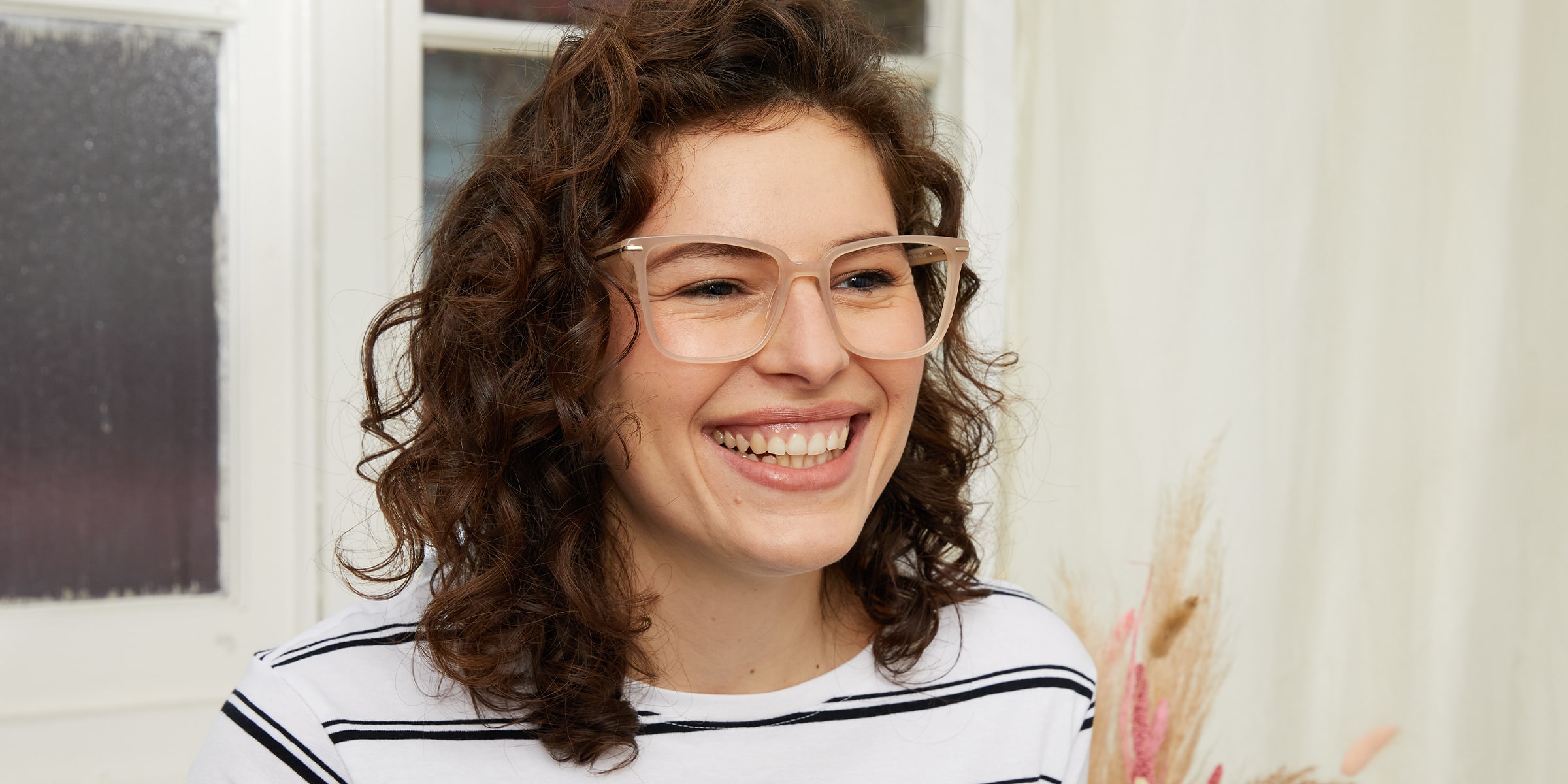 Smiling woman with curly brown hair wearing large, nude-coloured cat-eye glasses