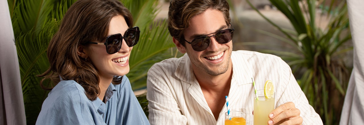 A laughing couple having drinks next to some palm fronds. The man wears cool aviator shades and the woman wears large butterfly sunglasses.