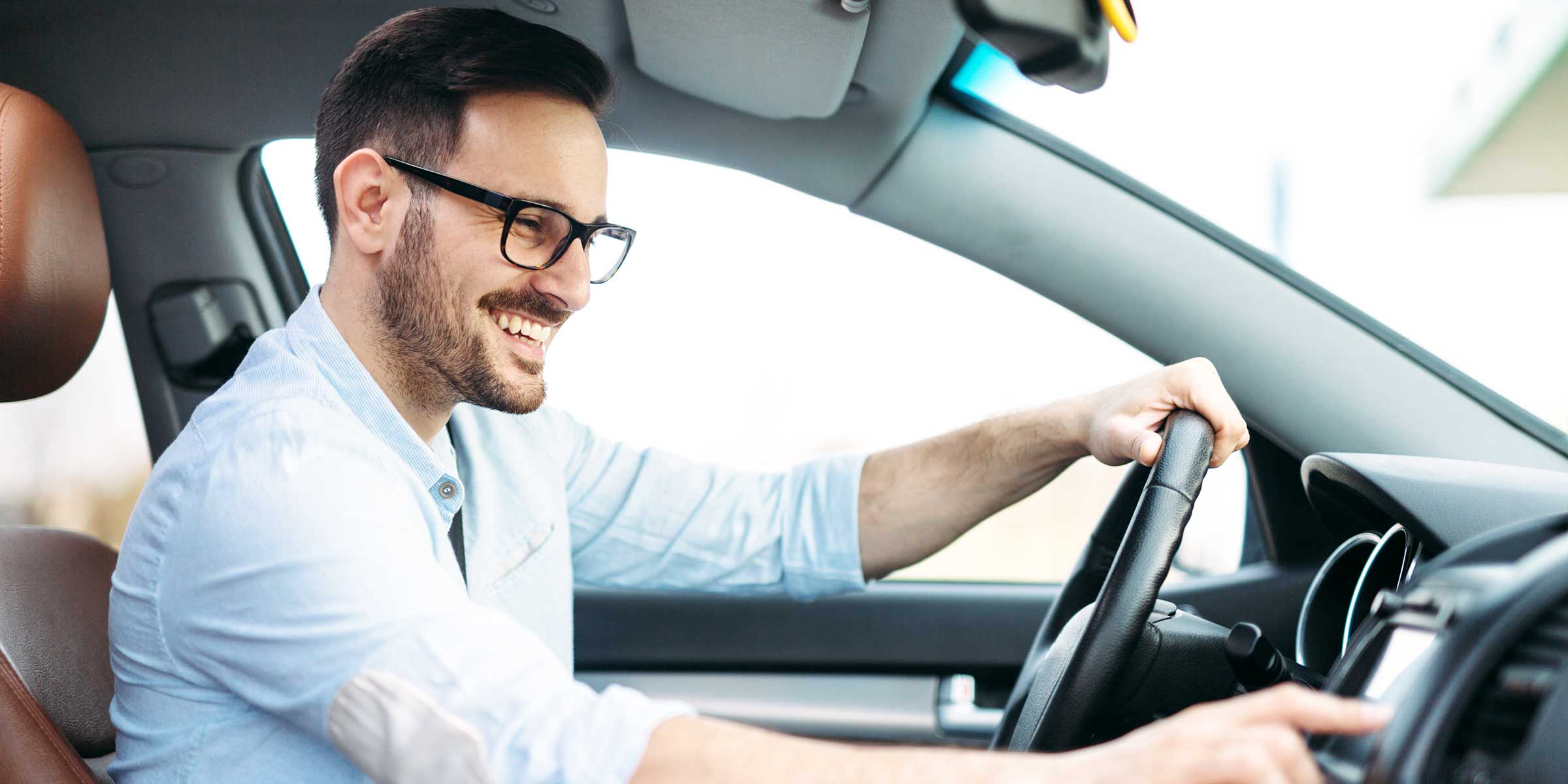 A smiling man with short hair and black rectangular glasses moves to adjust something on the dashboard in his car