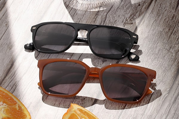 A black and a brown pair of sunglasses with grey lenses lying on a wooden table