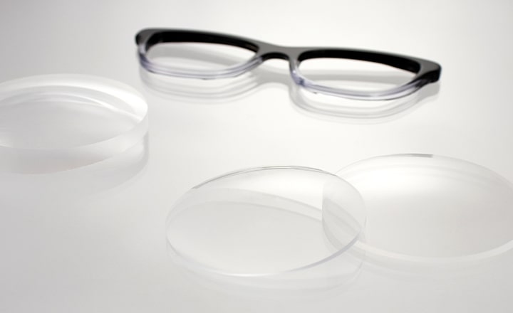 The front of a black-and-white glasses frame lying next to three uncut lenses on a table