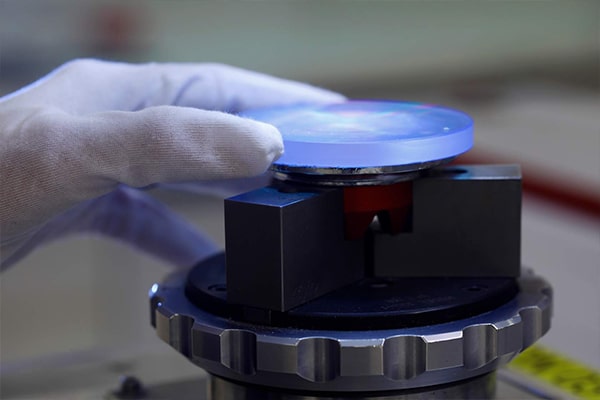 A gloved hand holding a glasses lens up against the work surface of a machine