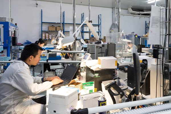 A cluttered warehouse space with several desks. Technicians at the desks are enterting data on their laptops.