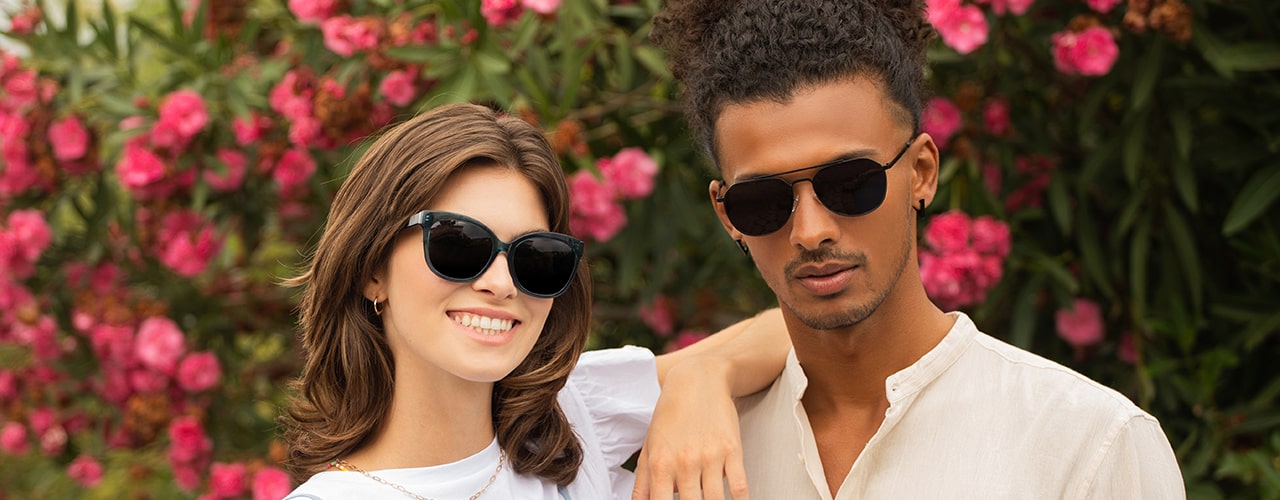 A couple posing in front of flower bushes. The smiling woman sports a pair of cat eye sunglasses, while the man wears a pair of cool aviator shades.