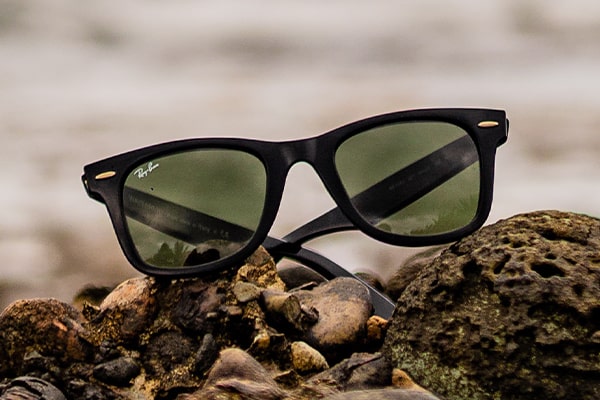 A black Ray-Ban wayfarer frame with green tinted lenses lying on a rocky beach